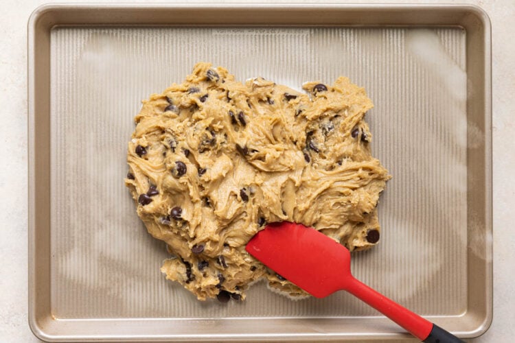 a red spatula spreading out cookie dough on a gold baking sheet.