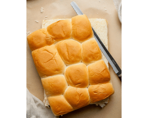 Overhead view of a loaf of Hawaiian rolls sliced in half horizontally with a serrated knife on parchment paper, ready to be assembled for pizza sliders.