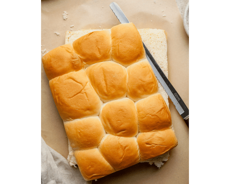 Overhead view of a loaf of Hawaiian rolls sliced in half horizontally with a serrated knife on parchment paper, ready to be assembled for pizza sliders.