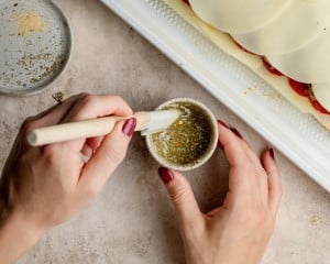 Close-up of hands mixing melted butter with Italian seasoning and garlic powder in a small bowl using a pastry brush, next to the assembled unbaked sliders.
