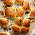 Overhead view of freshly baked pizza sliders being pulled apart, showing gooey melted cheese stretching between the rolls. The golden tops are brushed with garlic butter and sprinkled with Italian herbs and parsley, with bowls of marinara sauce nearby for dipping.