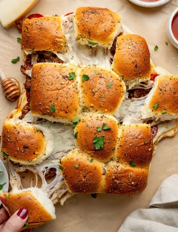 Overhead view of freshly baked pizza sliders being pulled apart, showing gooey melted cheese stretching between the rolls. The golden tops are brushed with garlic butter and sprinkled with Italian herbs and parsley, with bowls of marinara sauce nearby for dipping.