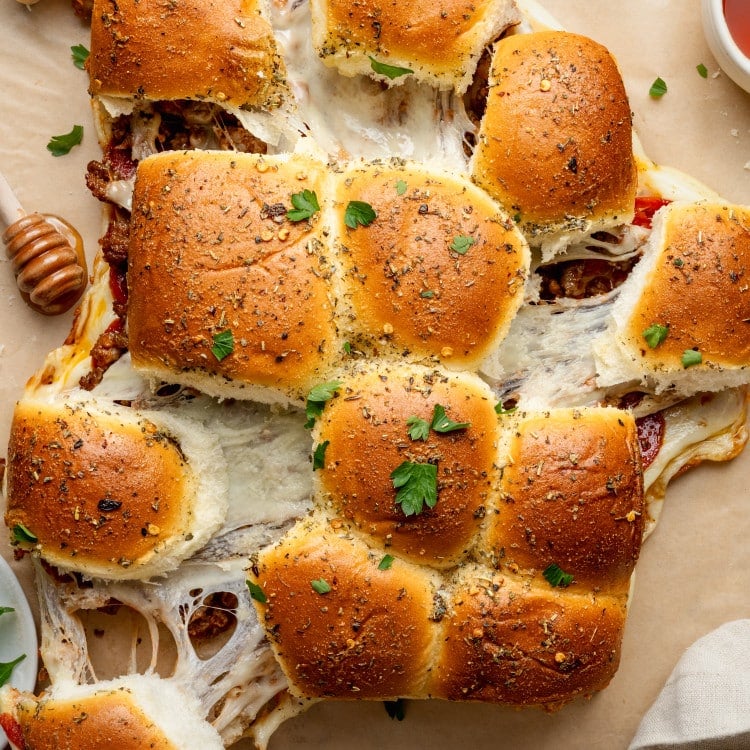 Overhead view of freshly baked pizza sliders being pulled apart, showing gooey melted cheese stretching between the rolls. The golden tops are brushed with garlic butter and sprinkled with Italian herbs and parsley, with bowls of marinara sauce nearby for dipping.