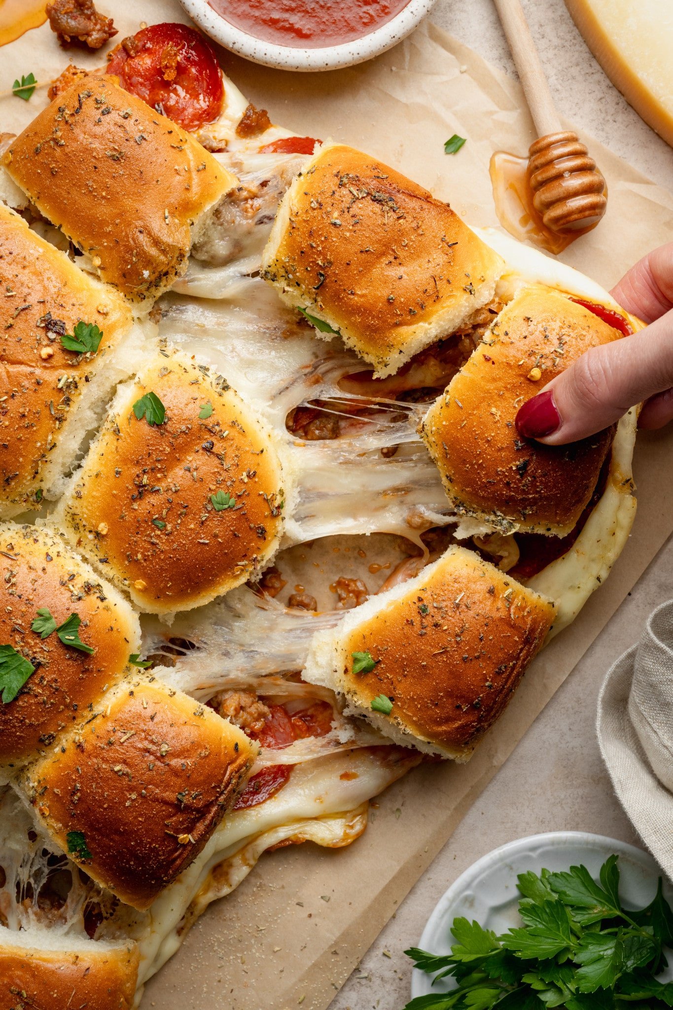 A hand pulls apart a cheesy pizza slider, showing gooey melted mozzarella stretching between the rolls. The sliders are golden brown with pepperoni and sausage layers visible inside.