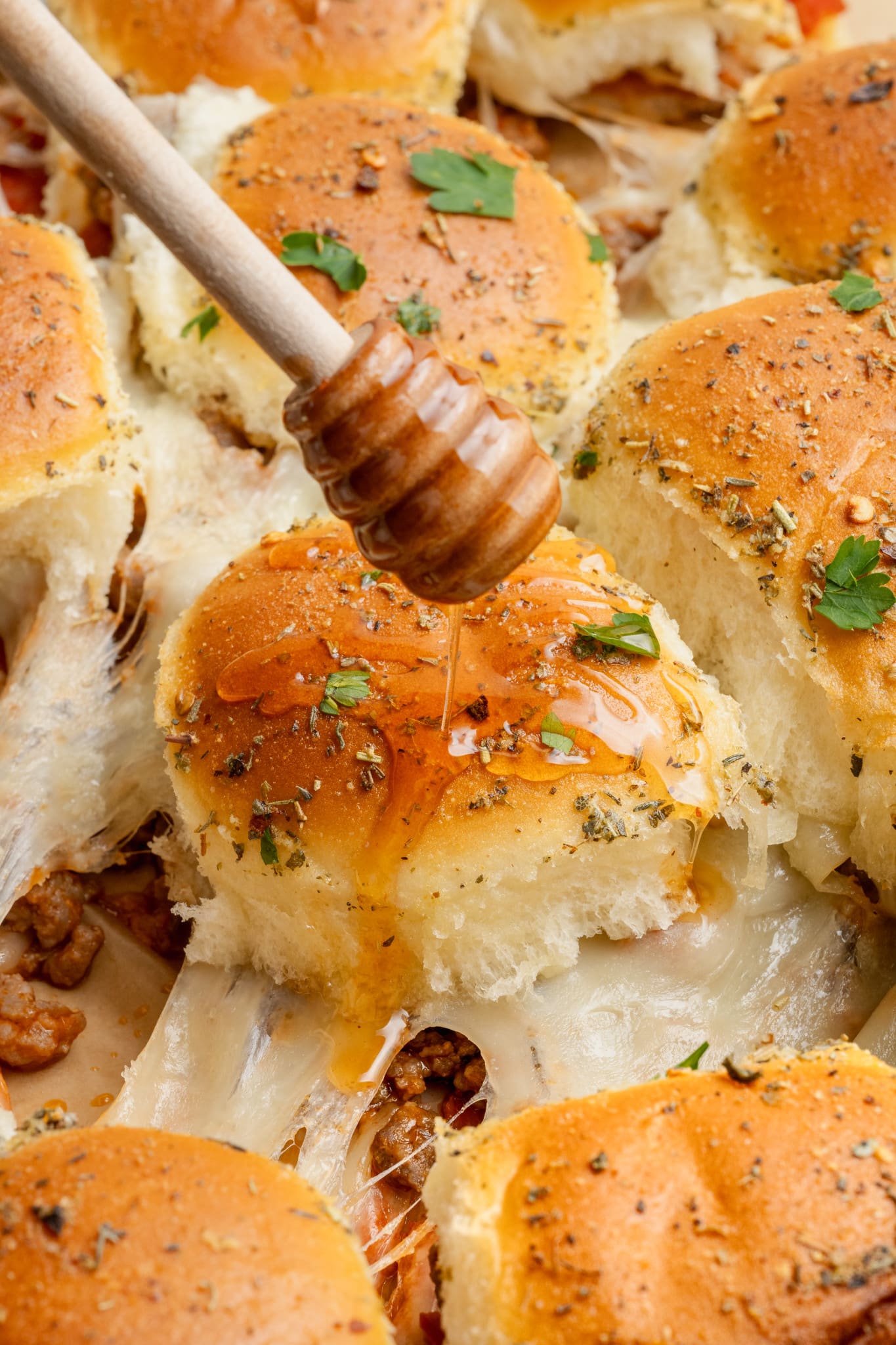 Close-up of pizza sliders being drizzled with honey from a wooden honey dipper. The rolls are golden, seasoned with herbs, and filled with melted cheese, sausage, and pepperoni.