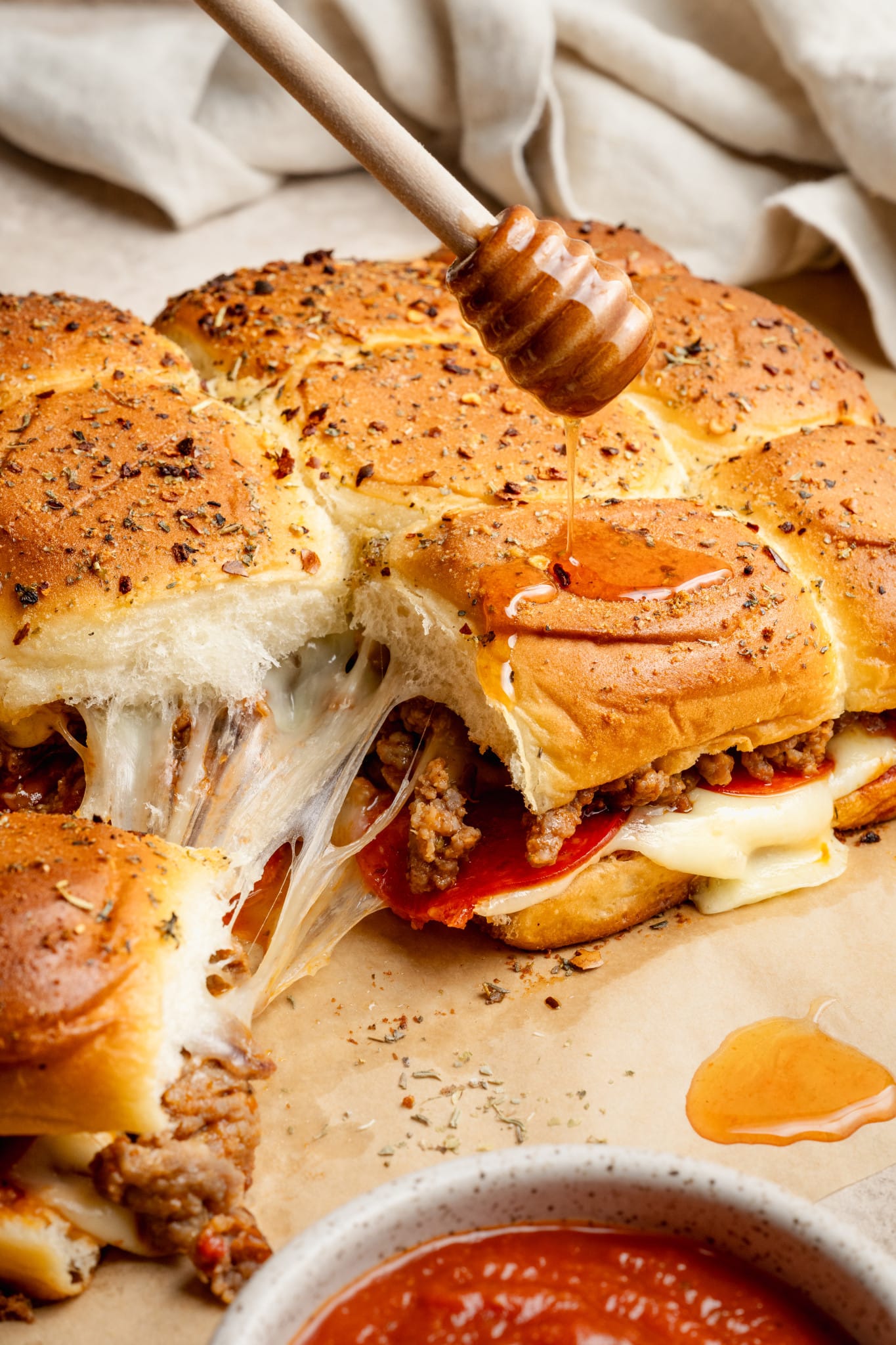 Close-up of honey being drizzled over hot pizza sliders with melted cheese stretching between the rolls. The tops are golden brown and sprinkled with crushed red pepper and herbs.