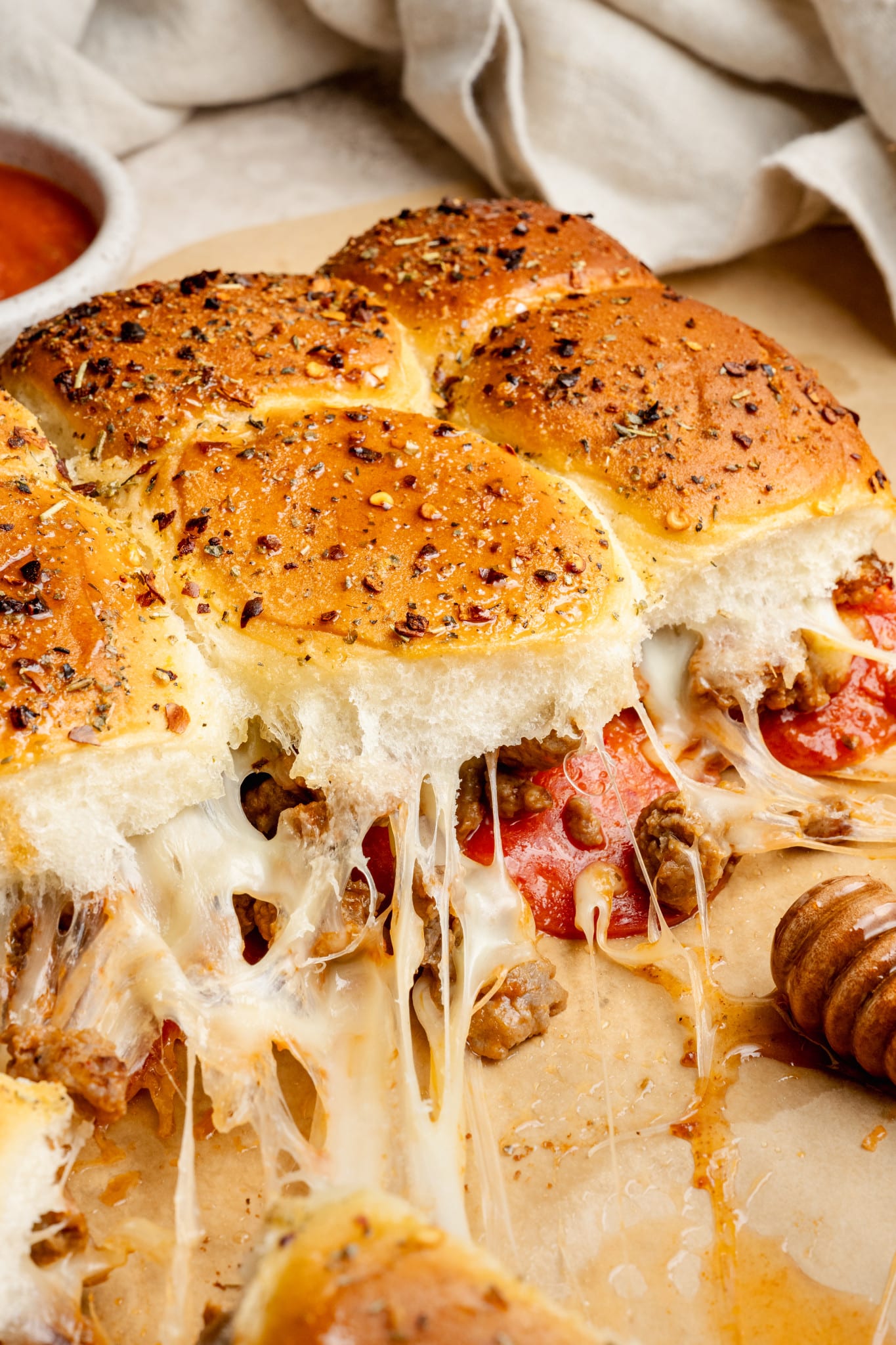 Close-up view of cheesy pepperoni and sausage pizza sliders, showing melted mozzarella pulling apart as the rolls are served. The golden tops are sprinkled with herbs and red pepper flakes.