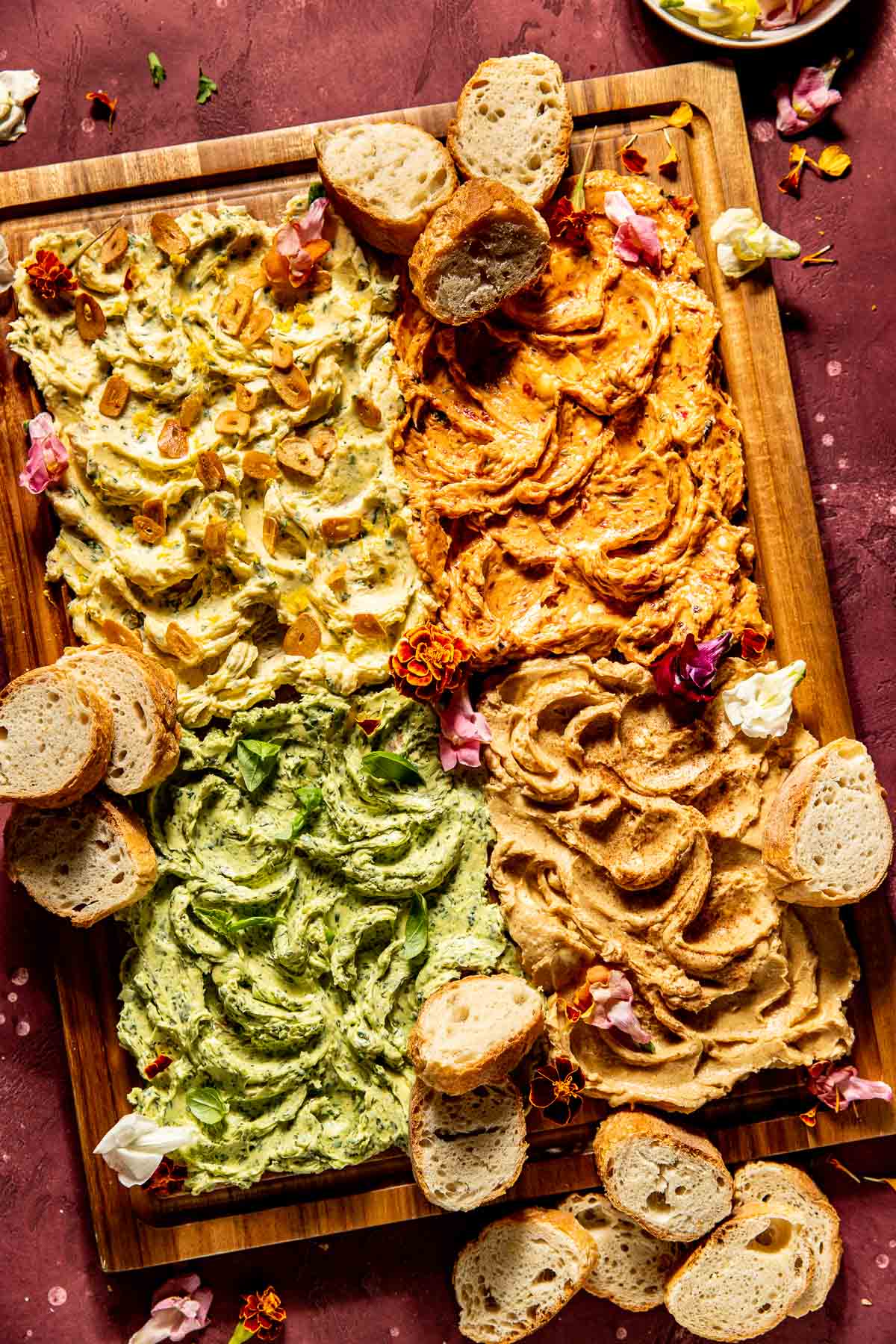 4 flavors of compound butters spread out on a wooden cutting board decorated with edible flowers and bread. 