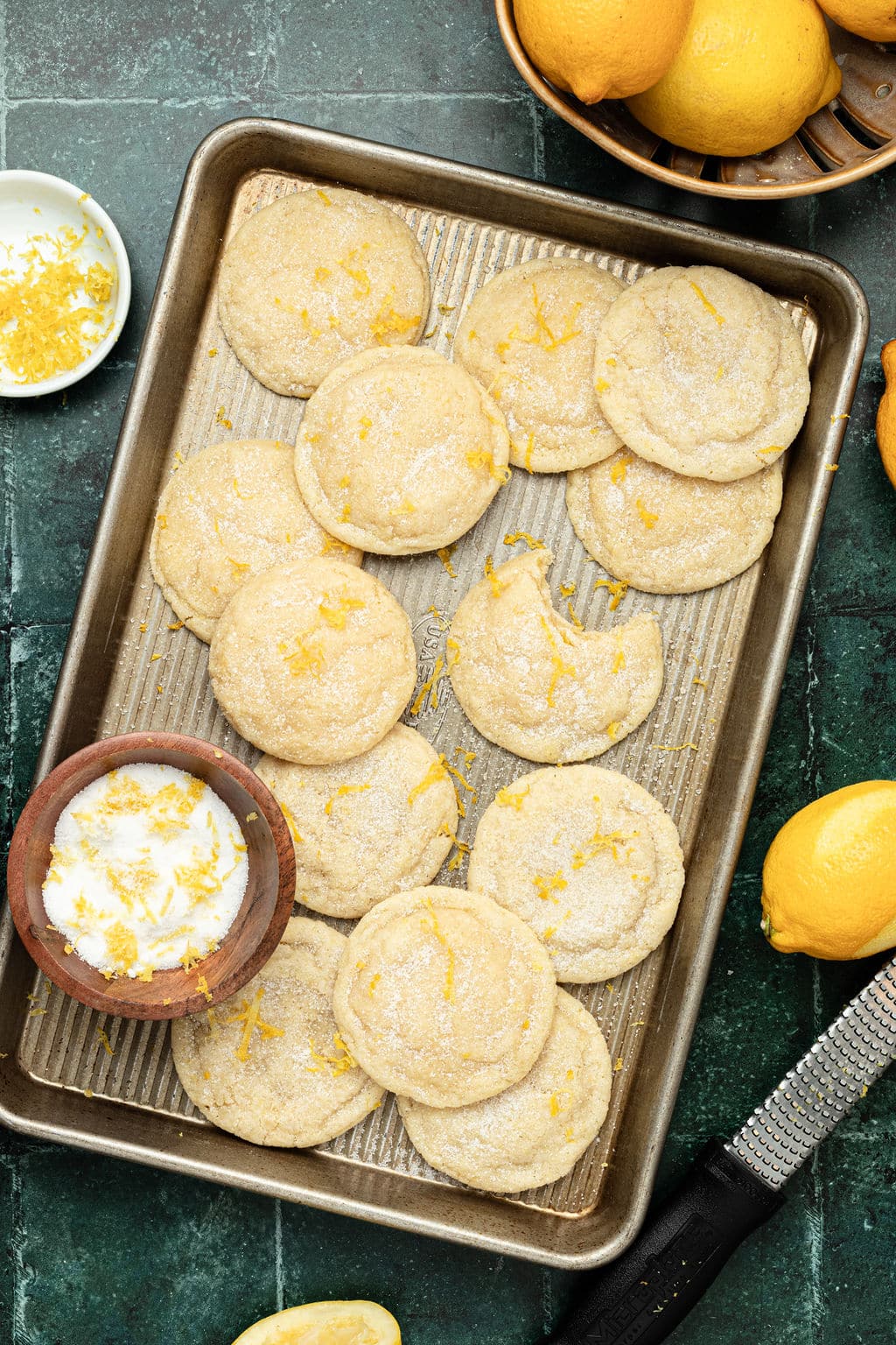 gold tray of sugar cookies topped with lemon zest plus a wooden bowl of sugar and lemon zeset, and lemons in the background