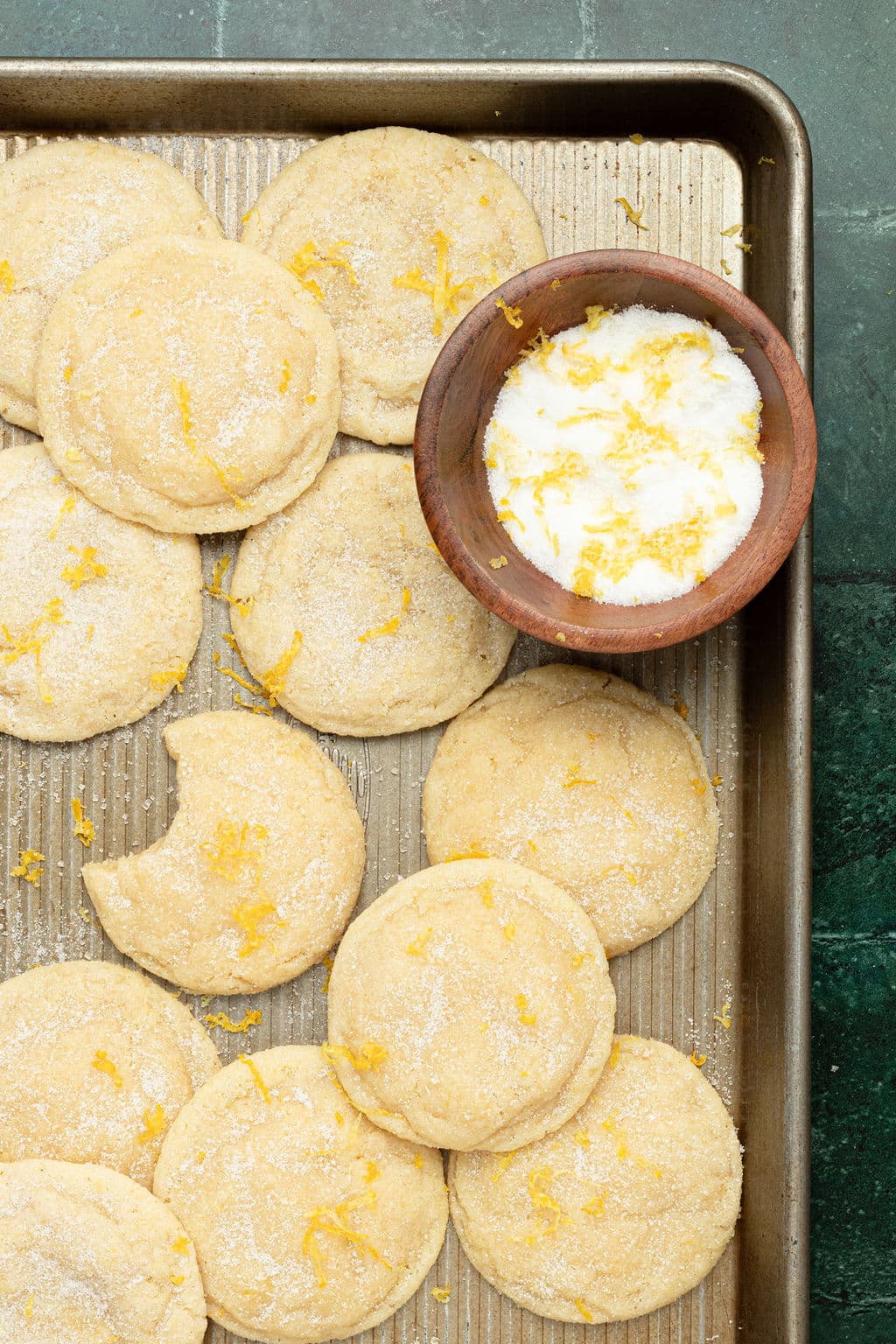 gold cookie sheet full of sugar cookies topped with lemon zest and sugar plus a small wooden bowl filled with sugar and lemon zest