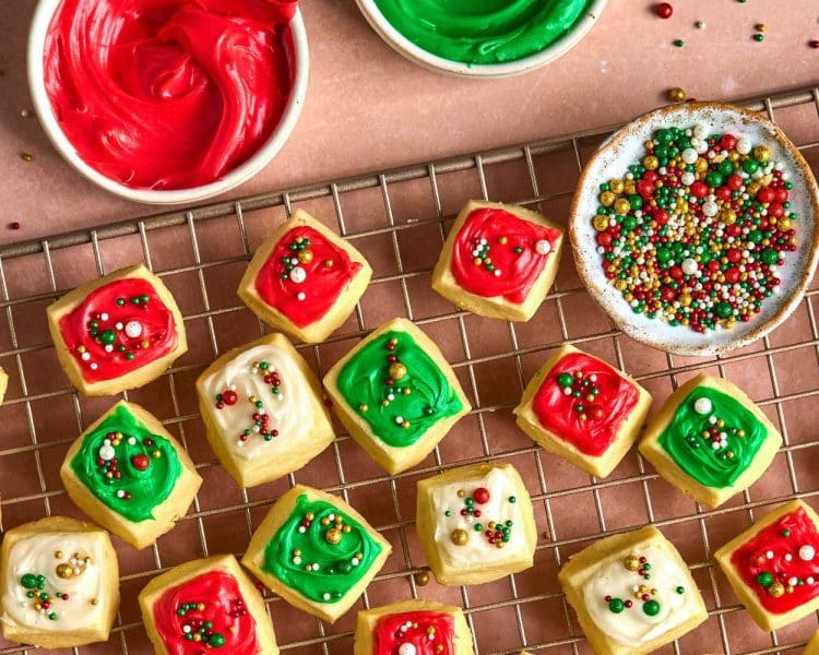 small square cookies with red, white, and green frosting and sprinkles, on a gold cooling rack. bowl of sprinkles and partial bowls of frosting also pictured