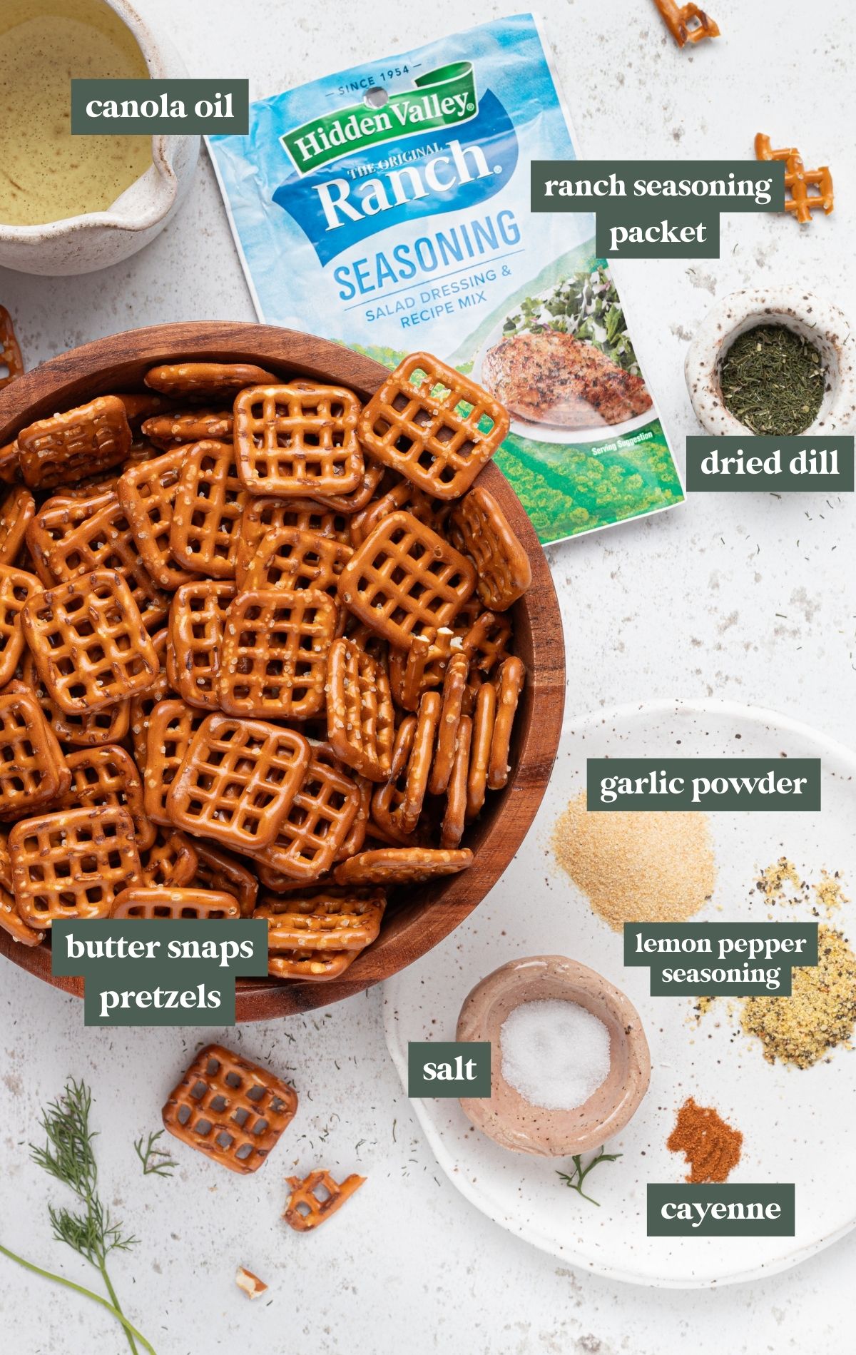 wooden bowl with square pretzels, small pitcher with oil, hidden valley ranch seasoning packet, dried dill, salt, garlic powder, lemon pepper seasoning, and cayenne