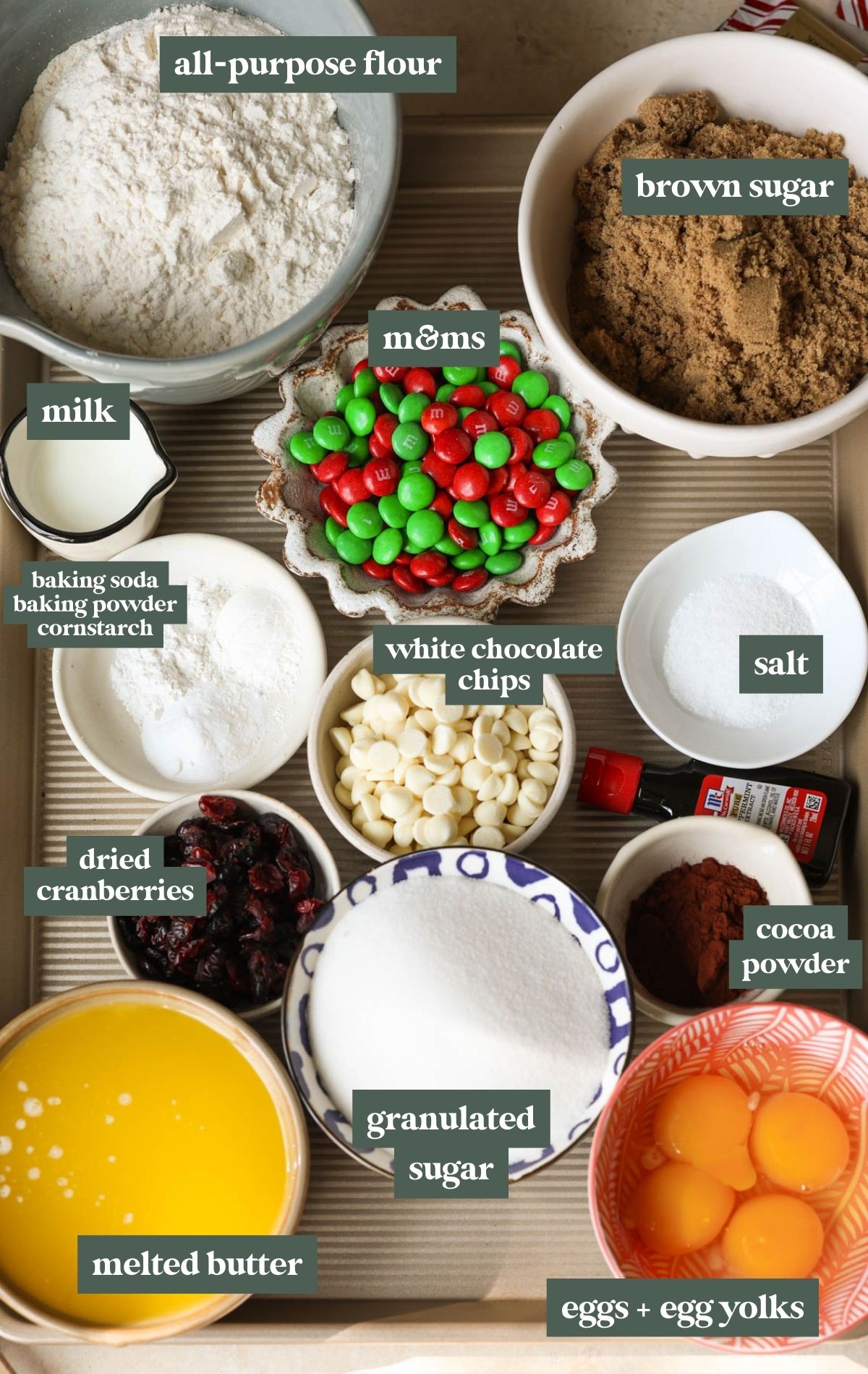 bowls with cookie ingredients including all purpose flour, brown sugar, milk, m&ms, baking soda, baking powder, cornstarch, white chocolate chips, salt, dried cranberries, cocoa powder, granulated sugar, melted butter, eggs, and egg yolks