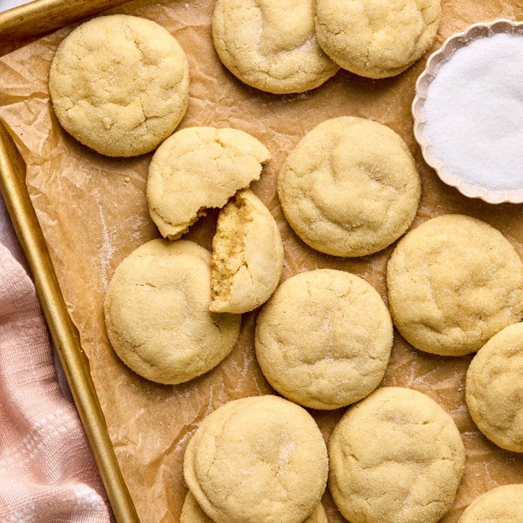 pile of sugar cookies on cookie sheet lined with parchment paper including one cookie split in half and a bowl with white granulated sugar