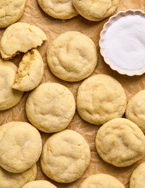 close up shot of pile of sugar cookies on cookie sheet lined with parchment paper including one cookie split in half and a bowl with white granulated sugar