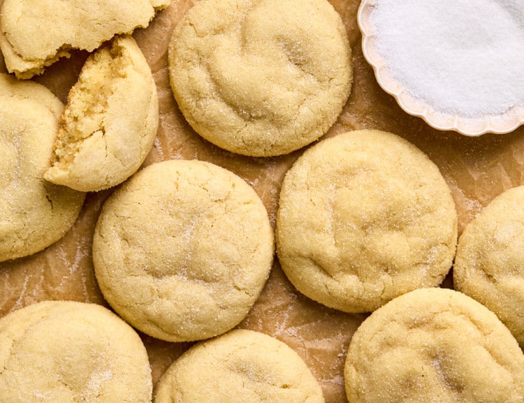 close up shot of pile of sugar cookies on cookie sheet lined with parchment paper including one cookie split in half and a bowl with white granulated sugar