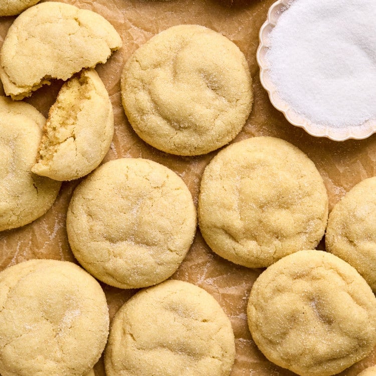 close up shot of pile of sugar cookies on cookie sheet lined with parchment paper including one cookie split in half and a bowl with white granulated sugar