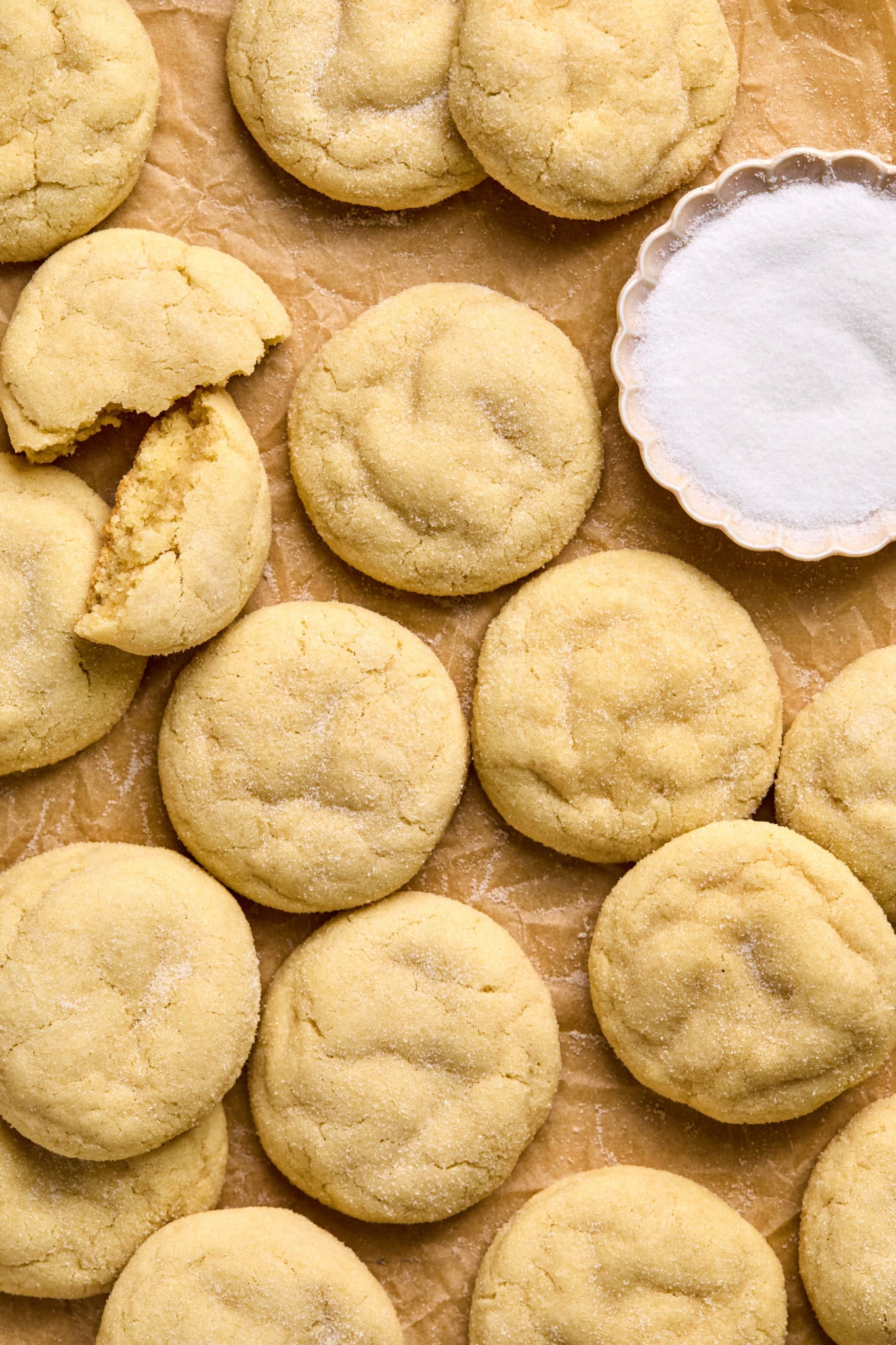 close up shot of pile of sugar cookies on cookie sheet lined with parchment paper including one cookie split in half and a bowl with white granulated sugar