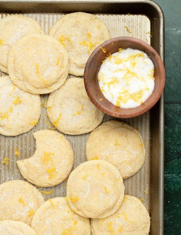 gold cookie sheet full of sugar cookies topped with lemon zest and sugar plus a small wooden bowl filled with sugar and lemon zest