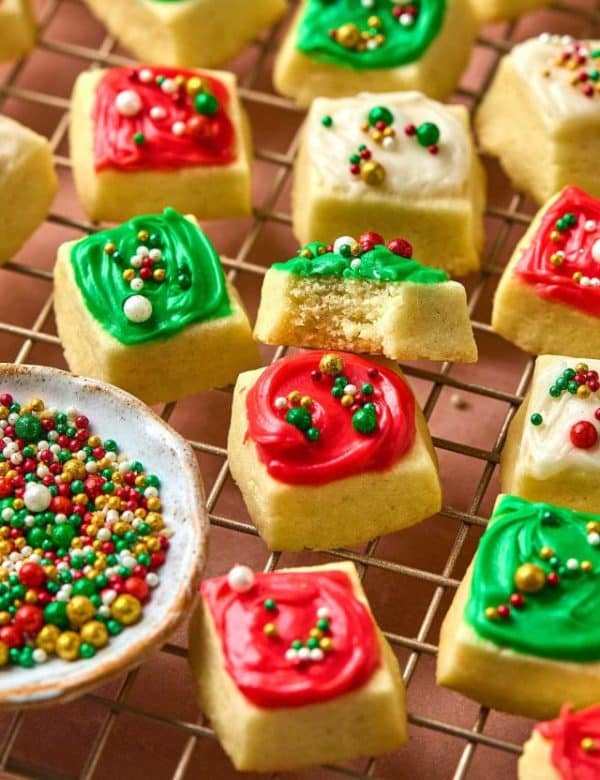 close up photo of small square cookies with red, white, and green frosting and sprinkles, on a gold cooling rack.