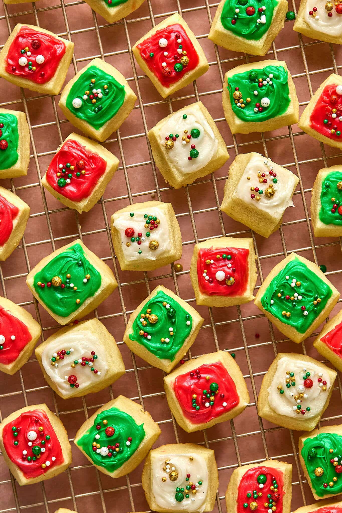 close up image of small square cookies with red, white, and green frosting and sprinkles, on a gold cooling rack.