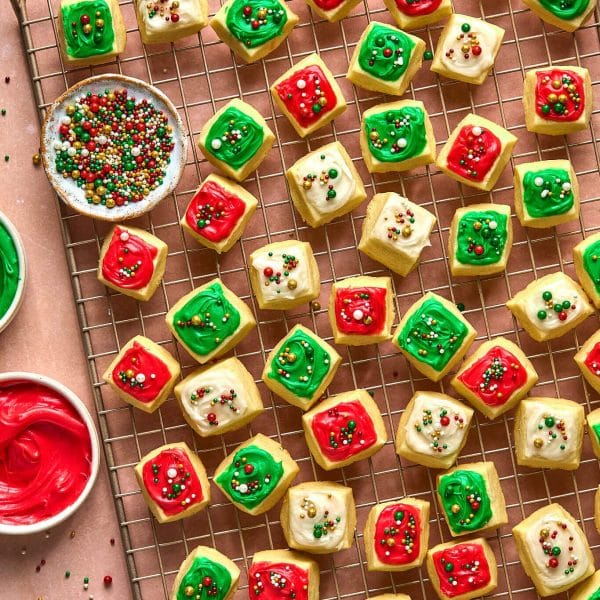 small square cookies with red, white, and green frosting and sprinkles, on a gold cooling rack. bowl of sprinkles and partial bowls of frosting also pictured