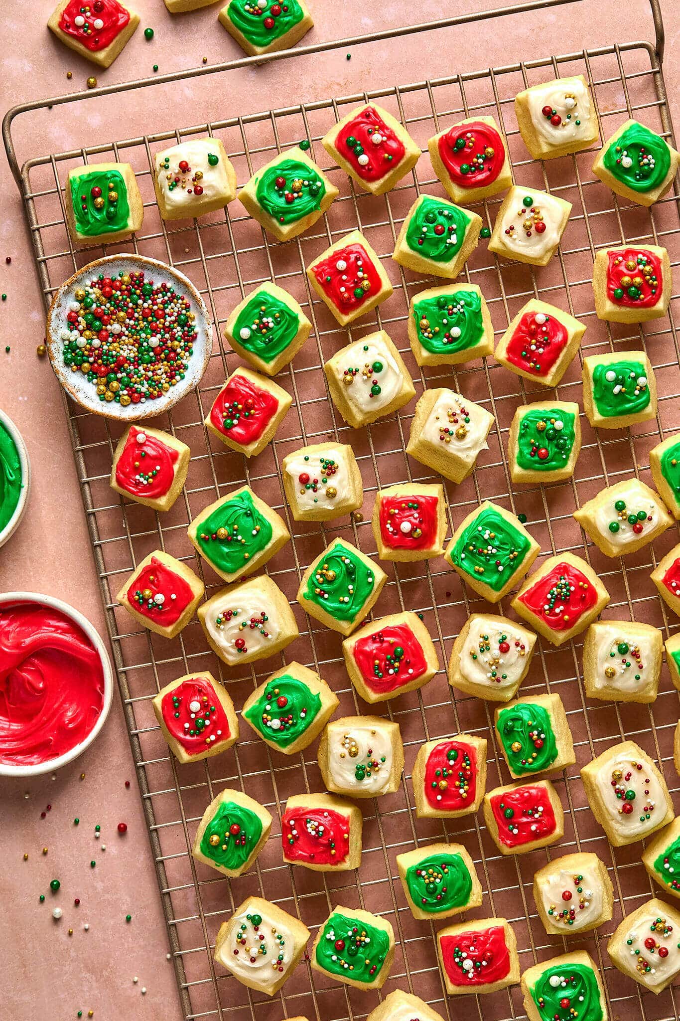 small square cookies with red, white, and green frosting and sprinkles, on a gold cooling rack. bowl of sprinkles and partial bowls of frosting also pictured