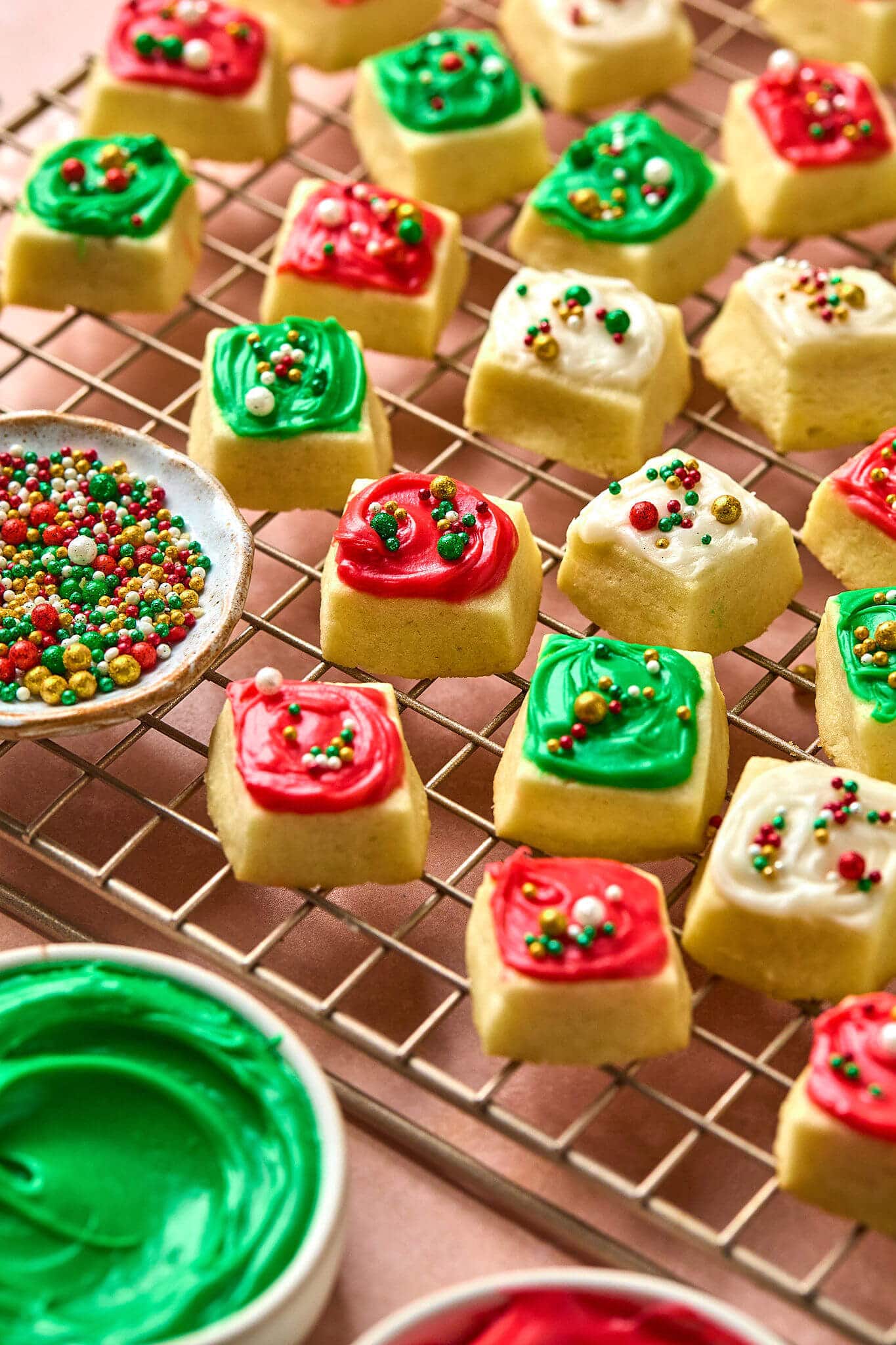 close up photo of small square cookies with red, white, and green frosting and sprinkles, on a gold cooling rack.