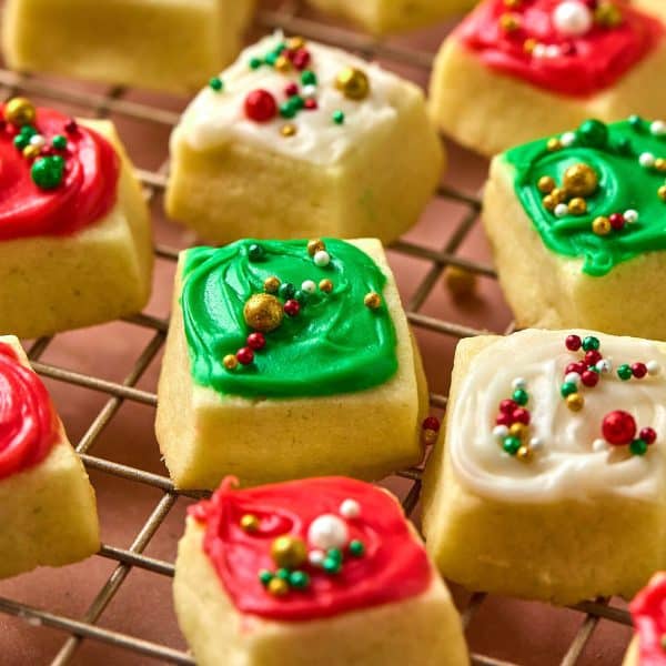 close up photo of small square cookies with red, white, and green frosting and sprinkles, on a gold cooling rack.