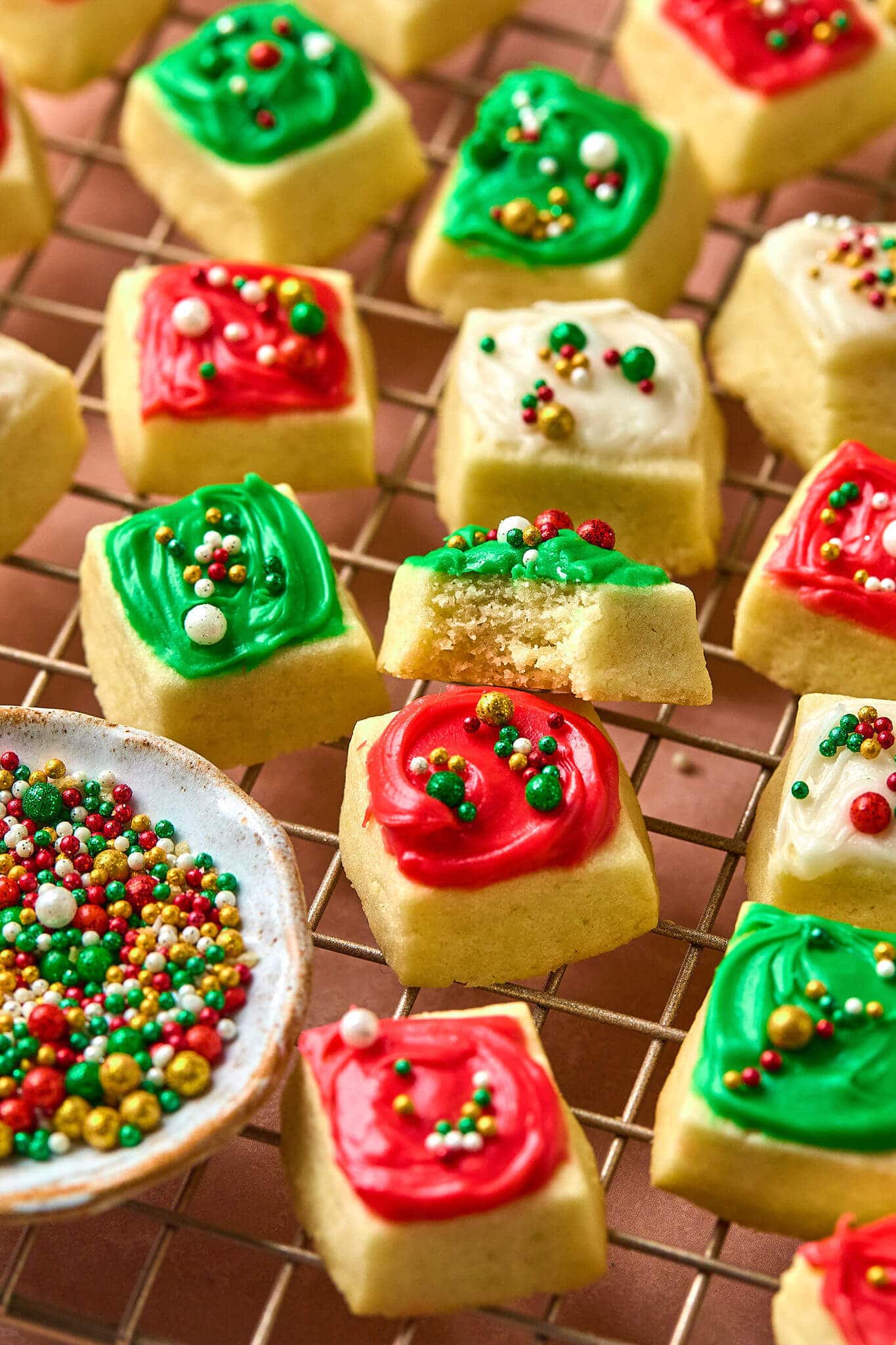 close up photo of small square cookies with red, white, and green frosting and sprinkles, on a gold cooling rack.