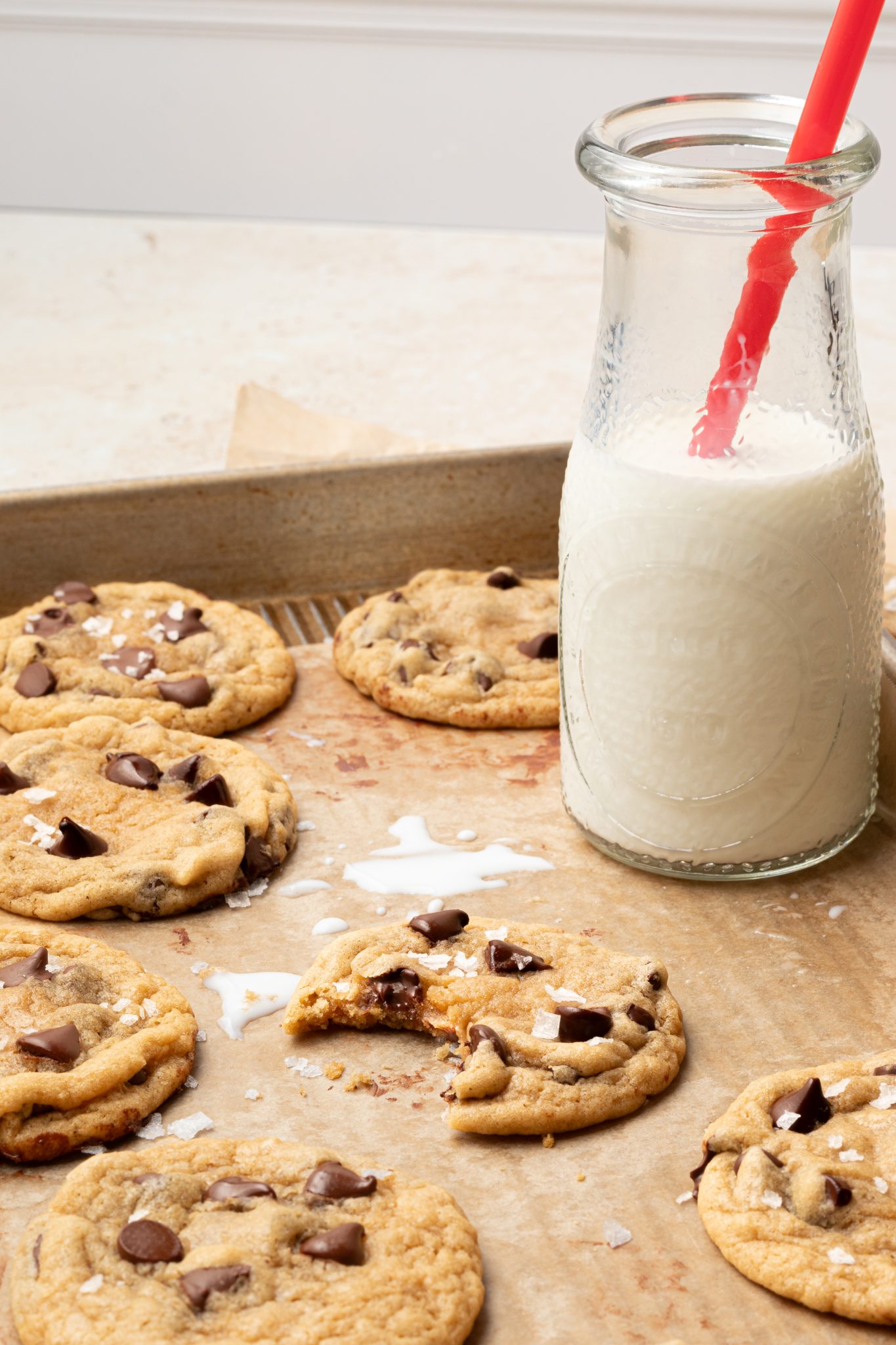 chocolate chip cookies on a parchment lined baking sheet with a glass of milk and a red straw. 