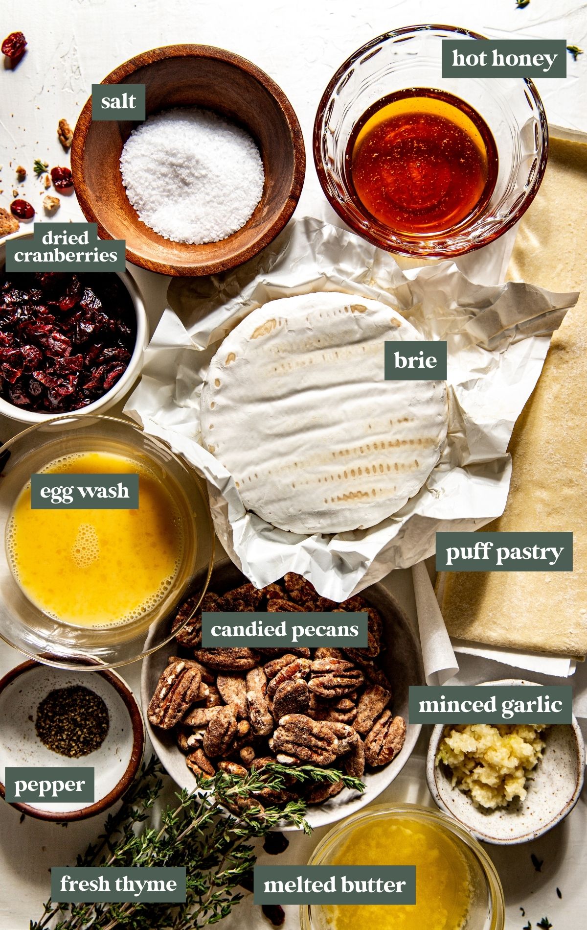 ingredients in multiple sized bowls including dried cranberries, hot honey, a wheel of brie cheese, egg wash, puff pastry, salt, pepper, candied pecans, minced garlic, melted butter, and fresh thyme.