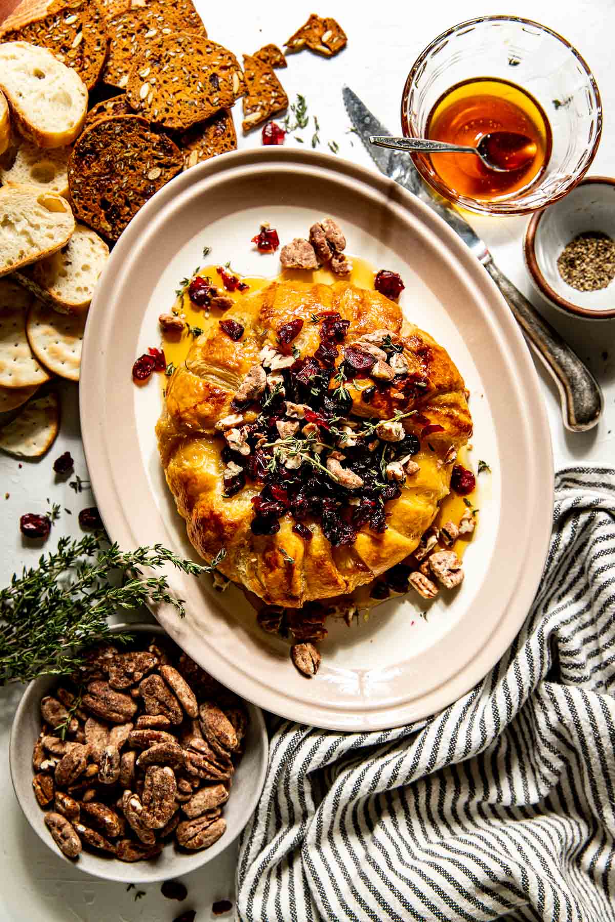 Round serving dish with a puff-pastry covered wheel of brie, topped with dried cranberries, fresh thyme, and candied pecans. Serving dish is surrounded by crackers, slices of baguette, and bowls of the toppings.