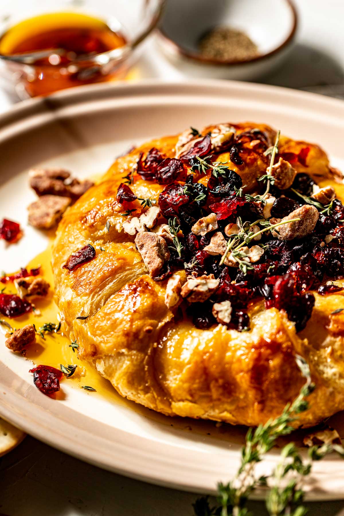 Close up image of a wheel of brie wrapped in puff pastry, topped with dried cranberries, candied pecans, and sprigs of fresh thyme.