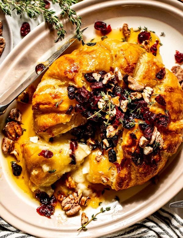 Round of brie cheese covered in puff pastry with a slice removed. Topped with dried cranberries, fresh thyme, and candied pecans.