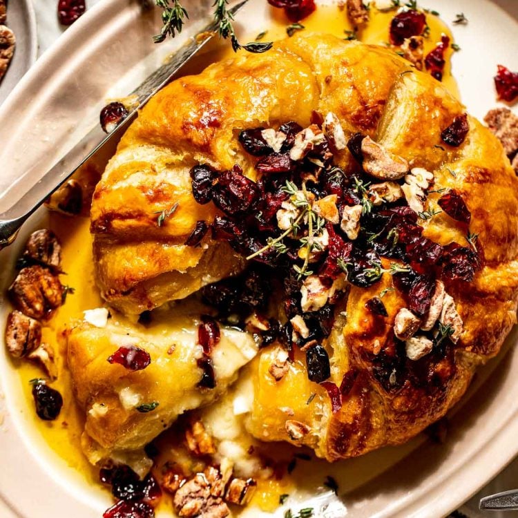 Round of brie cheese covered in puff pastry with a slice removed. Topped with dried cranberries, fresh thyme, and candied pecans.