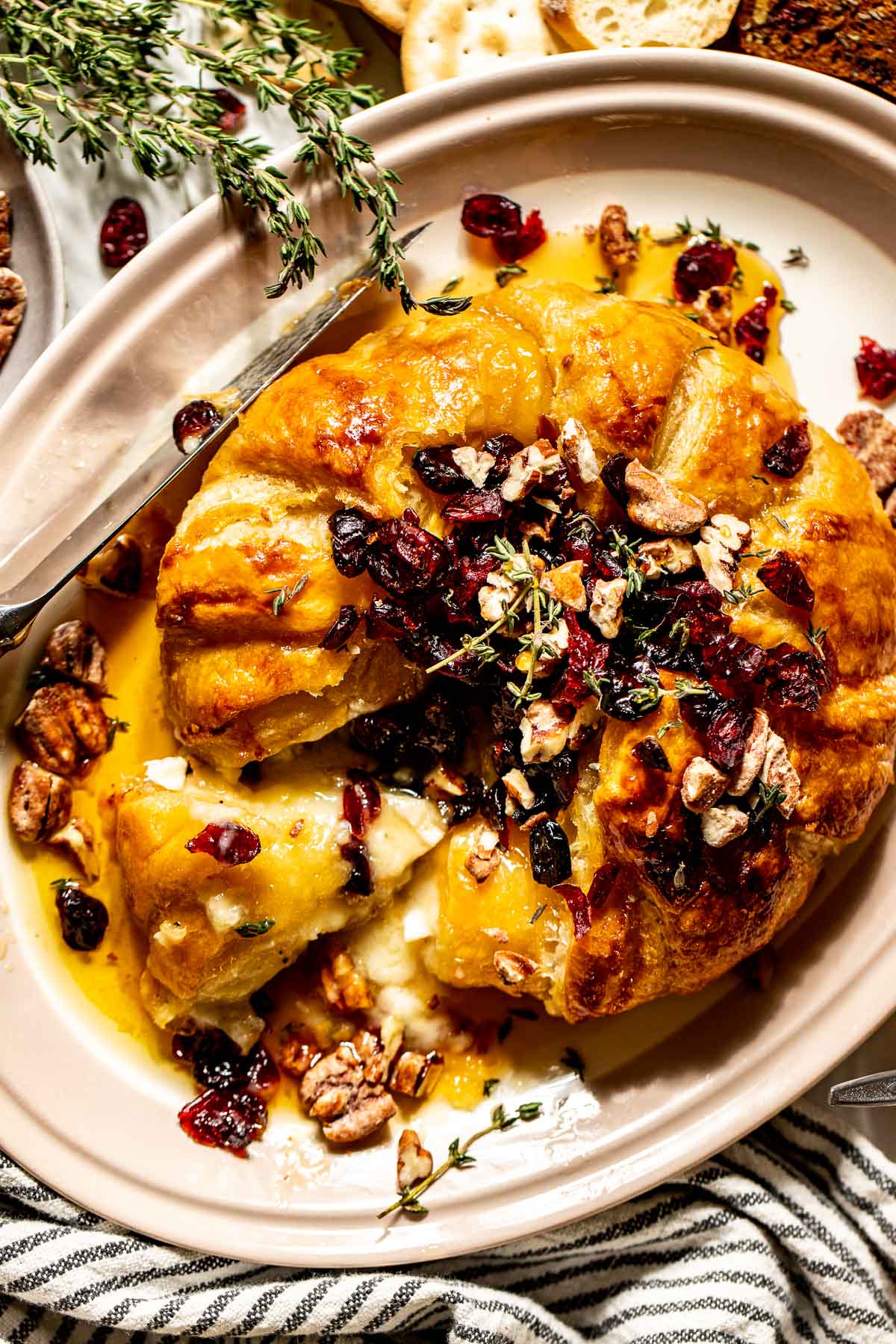 Round of brie cheese covered in puff pastry with a slice removed. Topped with dried cranberries, fresh thyme, and candied pecans.