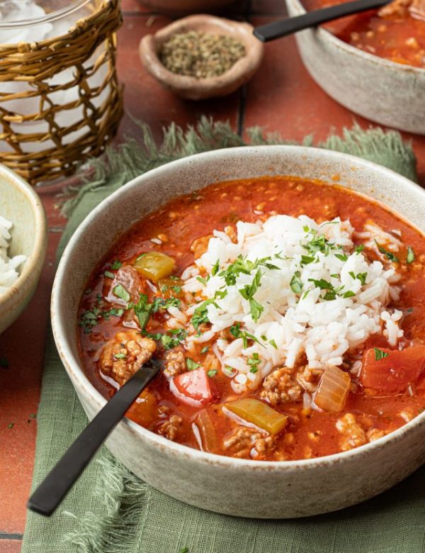 spoon in a bowl of soup with a red liquid base, chopped veggies and meat, topped with white rice and chopped parsley