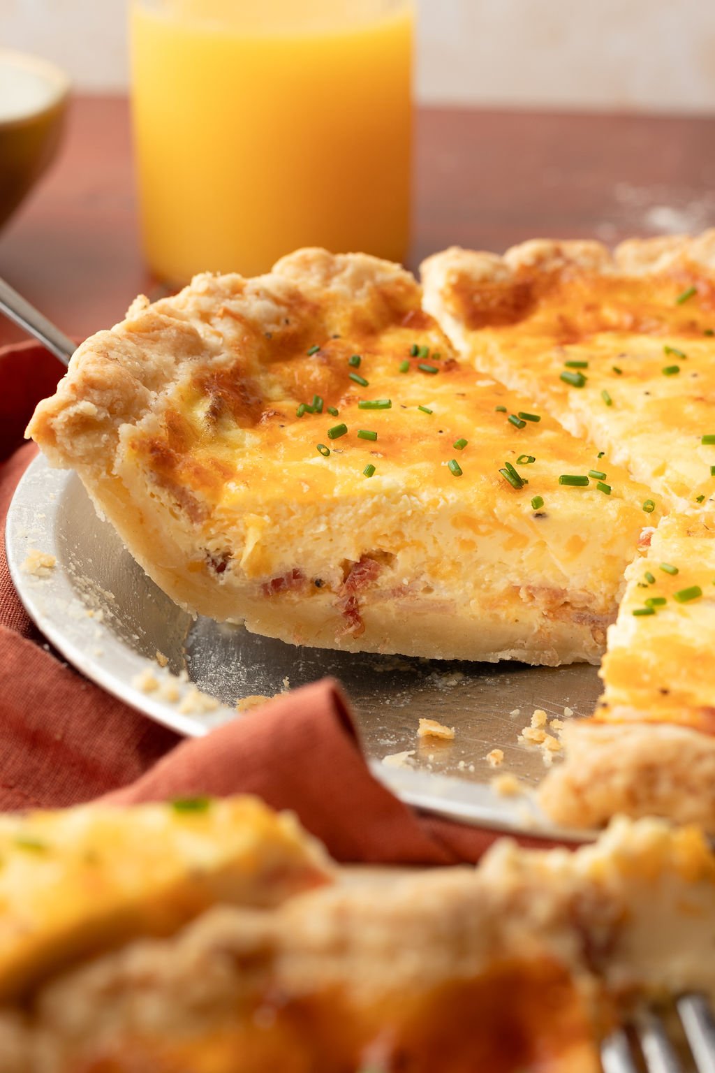 Up close image of a slice of quiche in a silver pie dish showing the egg center with bacon and topped with slices of green onion.