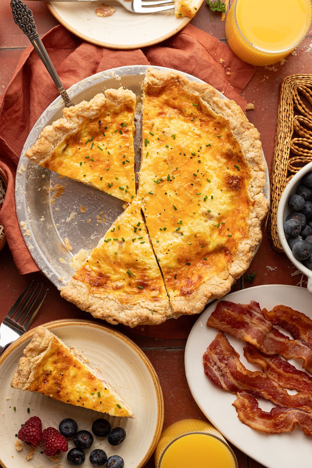 Full baked quiche with a slice missing, surrounded by plates with a slice of quiche, berries, bacon, and a glass of orange juice.