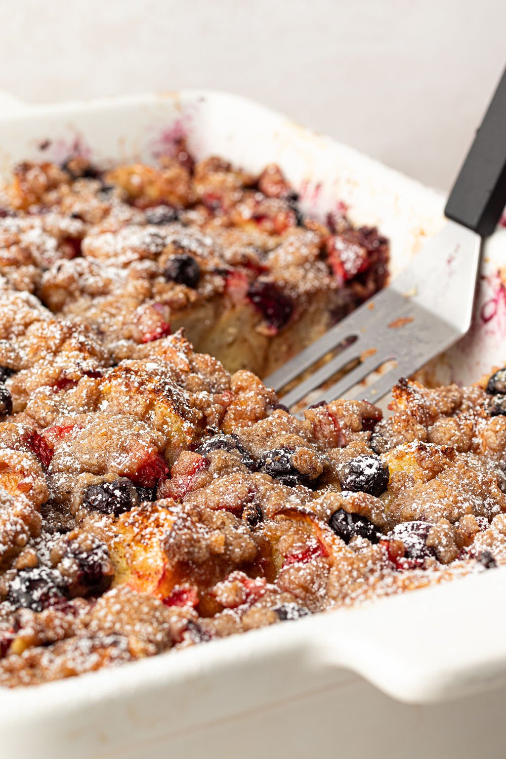 Offset spatula removing a piece of French toast with berries topped with cinnamon sugar and powdered sugar.