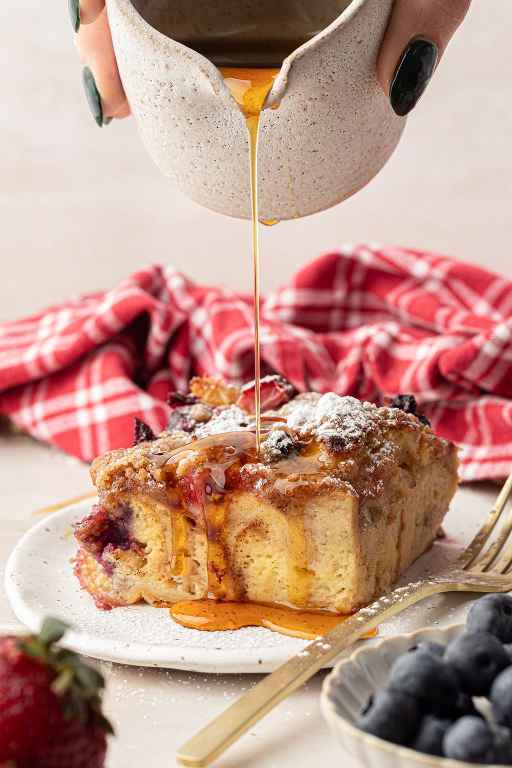 Syrup being poured over a slice of French Toast casserole with berries and powdered sugar.