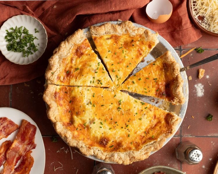 Whole baked quiche topped with green onion in a silver pie pan with small pieces missing.