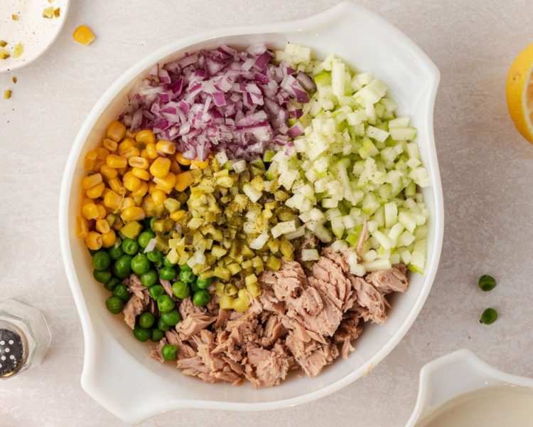 White bowl with corn, red onion, frozen peas, chopped gherkin pickles, tuna, and chopped green apple.