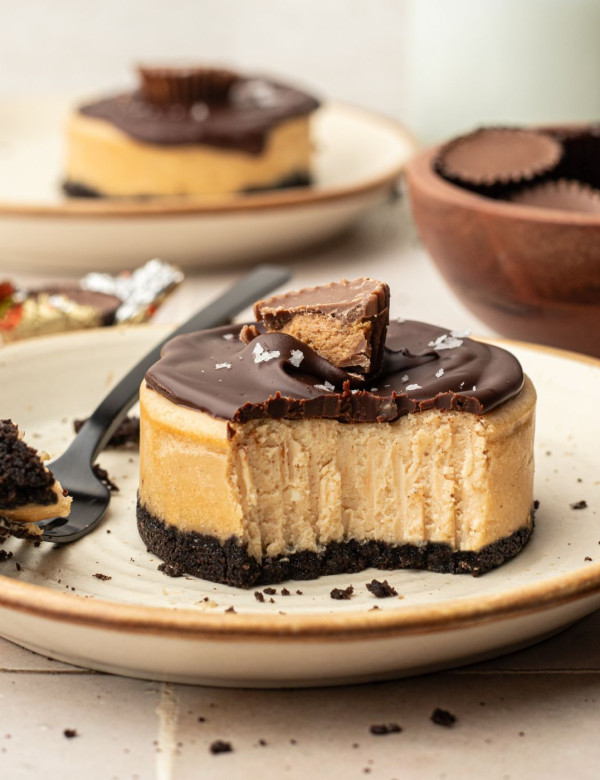 Plate with a circular cheesecake with peanut butter center, oreo cookie bottom, chocolate ganache topping, half of a reeses peanut butter cup, and flaky sea salt.