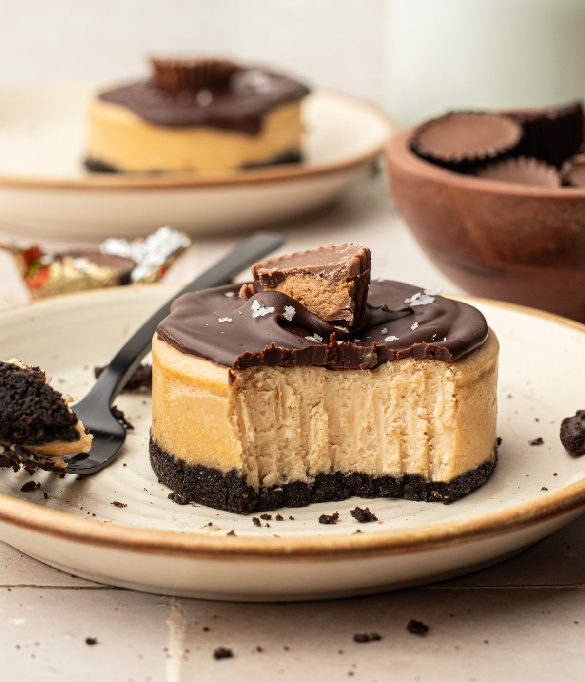 Plate with a circular cheesecake with peanut butter center, oreo cookie bottom, chocolate ganache topping, half of a reeses peanut butter cup, and flaky sea salt.