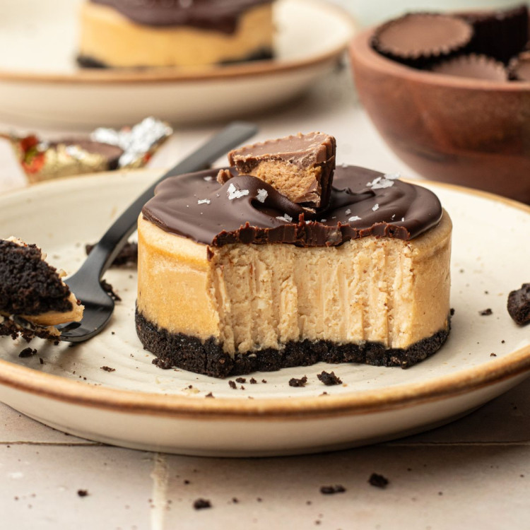 Plate with a circular cheesecake with peanut butter center, oreo cookie bottom, chocolate ganache topping, half of a reeses peanut butter cup, and flaky sea salt.