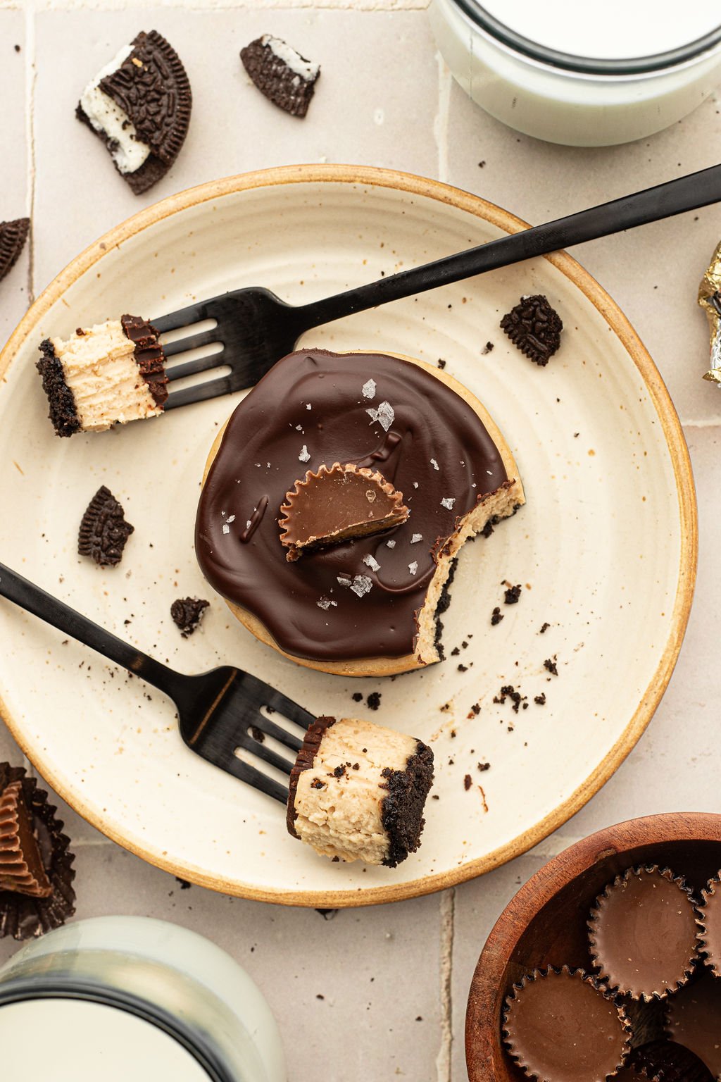 Overhead shot of a plate with a small circular cheesecake, topped with a chocolate ganache and half of a mini reeses cup. Two forks are on the top and bottom sides of the cheesecake, each with a bite on the fork.