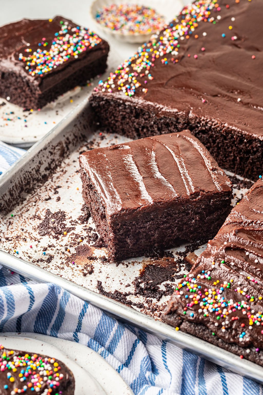 Square piece of chocolate cake with chocolate frosting on a sheet pan with the remainder of the cake with multi color sprinkles.