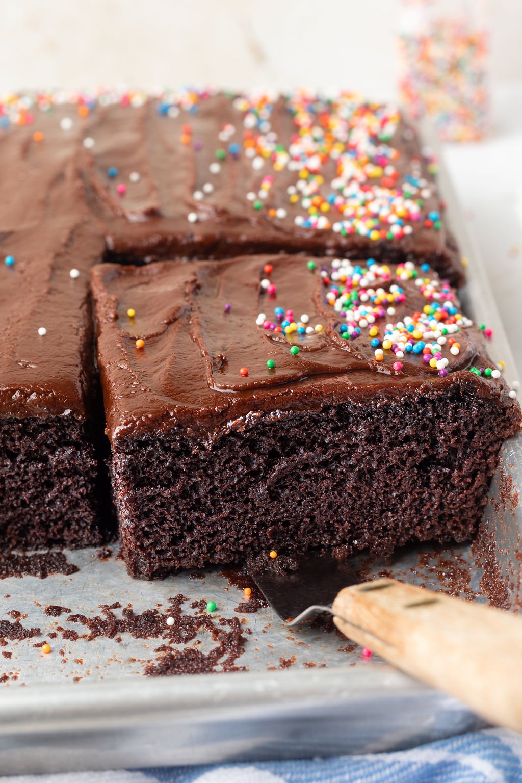 Square piece of chocolate cake with chocolate frosting and multi colored sprinkles being removed from sheet pan of cake.